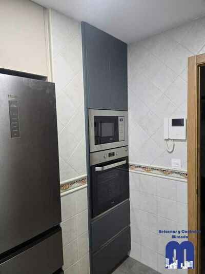 Reforma integral de cocina moderna con mobiliario en acabado tacto seda Fenix verde comodoro y beige, encimera y frente porcelánico Coverlam imitación mármol, suelo vinilico gris, fregadero bajo encimera y grifo monomando flexible de color negro.